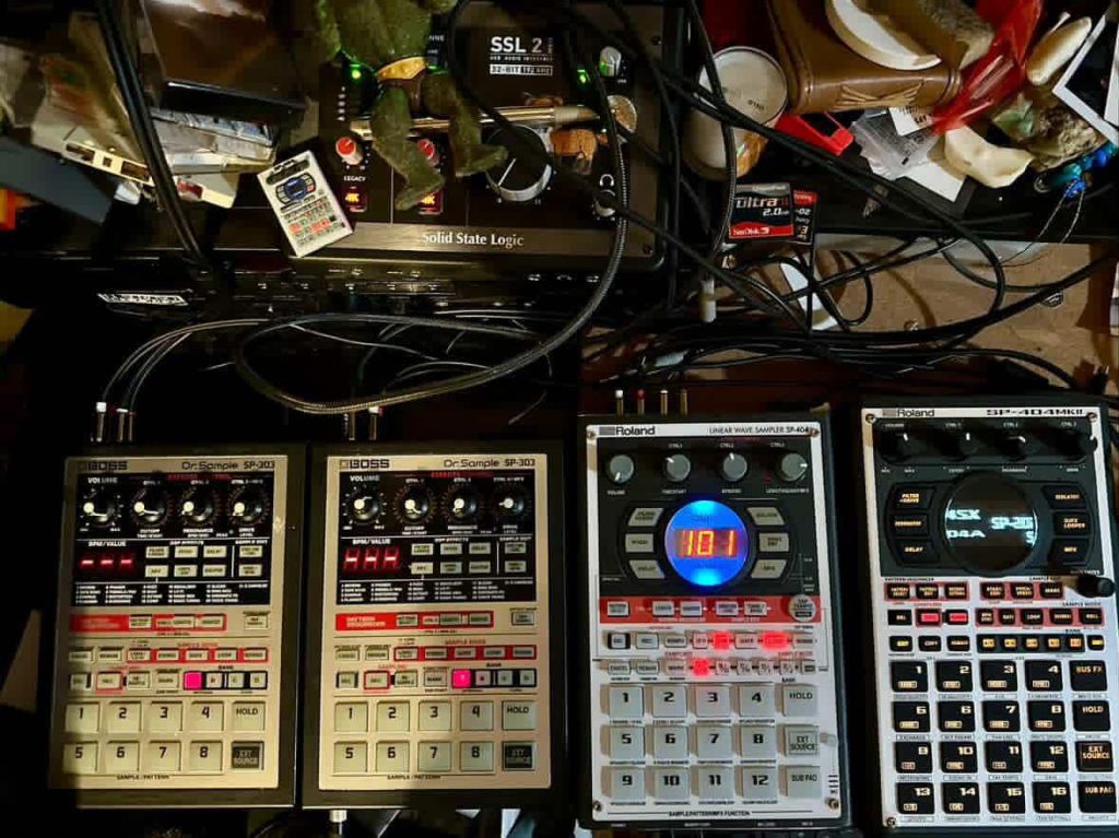 A cluttered tabletop view featuring multiple music production devices, including two Boss Dr Sample SP-303 units, a Roland SP-404 MKII, and an SP-404, along with connecting cables and a Solid State Logic interface.